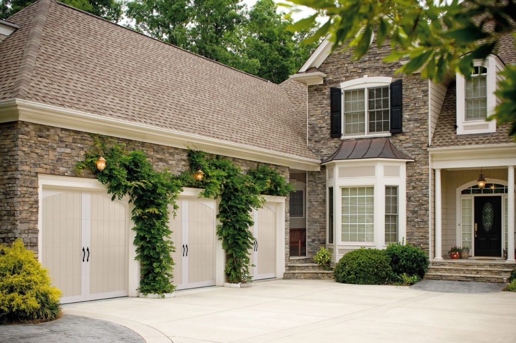 Large stone house with three-car garage, with vertical wood-panel white garage doors and vines on walls between the garage doors.