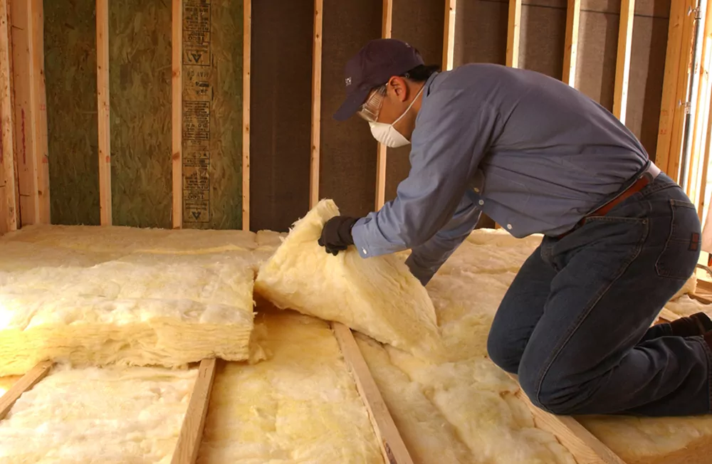 Technician kneeling and installing yellow batt insulation in an attic while wearing a face mask.