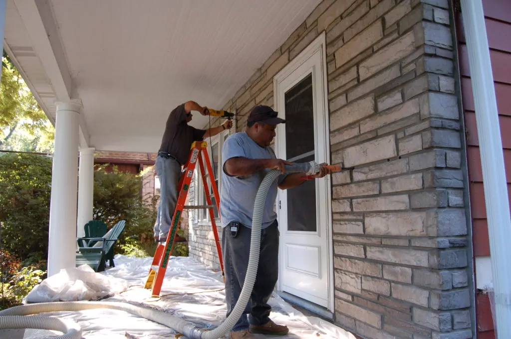 Technicians standing on home's porch, installing retrofit insulation through the front stone facade.